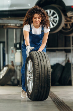Female auto mechanic with curly hair lifts a car tire in a garage, with vehicles and tools visible in the background, showcasing an automotive service environment