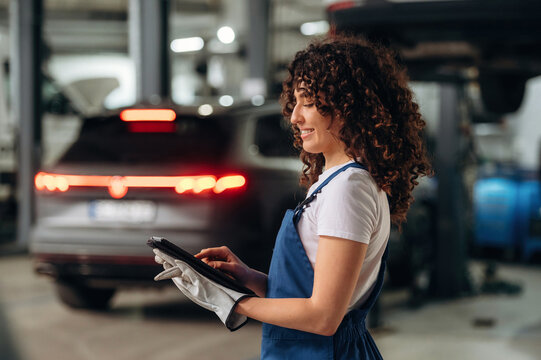 Female auto mechanic with curly hair using tablet in garage, modern car in background, wearing gloves and blue overalls, focused on work tasks and vehicle maintenance