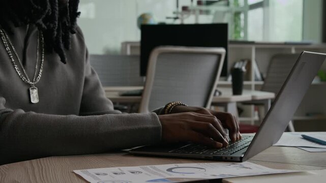 Close up view of Black man with dreadlocks working on laptop at desk with printed financial charts nearby during office day