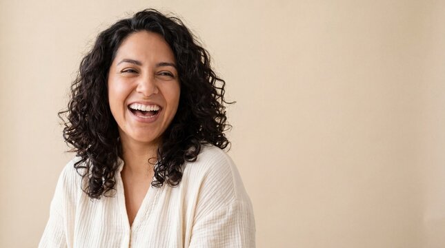 Smiling Hispanic woman with curly hair in neutral blouse, joyful expression and relaxed pose in studio portrait conveying warmth and confidence