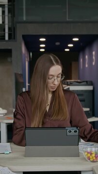 Vertical shot of young woman in glasses typing on digital tablet and checking documents at desk in modern open plan office