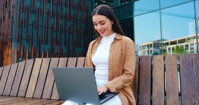 Happy young professional woman holding laptop and smiling while sitting on bench and looking to the side near a contemporary office building