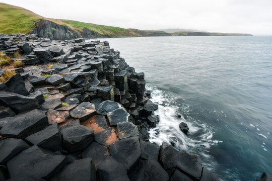 View of hexagonal basalt columns along the rugged coastline with ocean waves and green hills under an overcast sky Hofsos, Skagafjordur, Iceland.