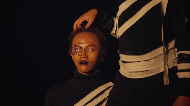 A still performer with spider makeup and white contacts is joined by another from above. A slow, creepy, and artistic interaction against a stark black background.