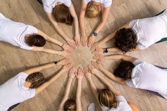 Diverse group of women in white shirts holding hands in circle on wooden floor.