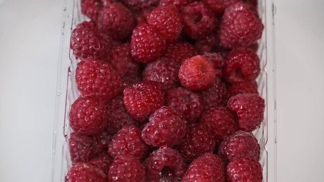 A close-up view displays a heap of fresh, ripe red raspberries sitting in a clear rectangular plastic punnet. Concept of healthy snacking, organic berry harvesting, and summer fruit nutrition