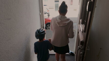 Fototapeta premium Mother opens door while holding toddler son with helmet as older child follows behind, family steps out of apartment together during morning departure