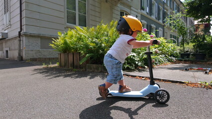 Fototapeta premium Toddler wearing helmet rides scooter outside residential building on sunny day, gliding along sidewalk near plants and apartment windows in suburban setting