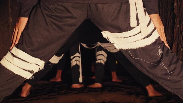 A unique low-angle perspective of a group of barefoot contemporary dancers in black and white costumes, standing in formation within a dark, earthy tunnel. The atmosphere is mysterious and tribal.