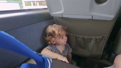 Toddler sits upright inside moving bus after crawling between seats, looking down and touching edge of seat during trip © Marco