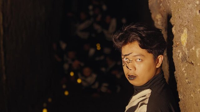 In a dark, mysterious tunnel, a man quickly turns from watching a group to stare directly at the camera, revealing his startling spider makeup and white contact lenses in a shocking moment.