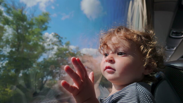 Toddler gazes through bus window with hand pressed to glass, face half-lit by sun and surrounded by blue sky and fluffy clouds reflected on surface