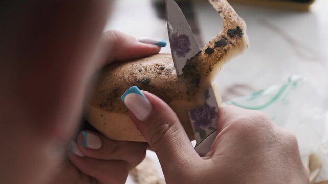 Over-the-shoulder close-up of a woman peeling an unwashed potato with a decorative knife. Focus on the domestic task of preparing fresh ingredients for a homemade meal.