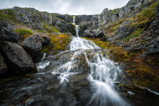 View of a multi-tiered waterfall cascading over dark volcanic rocks and vibrant green moss under an overcast sky in Eskifjordur, Fjardabyggd, Iceland.