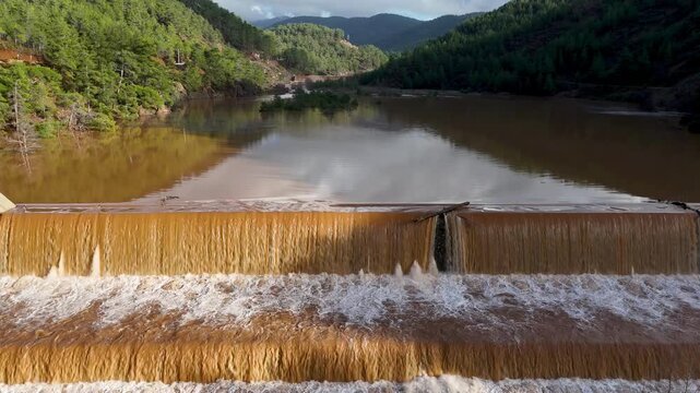 Muddy floodwaters of the Namnam River surging over a concrete stepped weir surrounded by pine-forested hills in Koycegiz, Mugla, Turkey. Aerial drone view with no movement