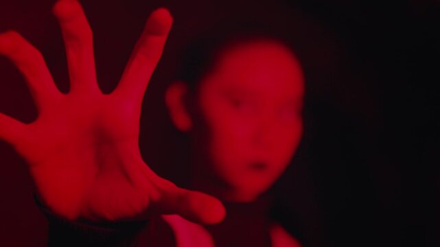 In dramatic red light, a woman with creepy white contact lenses is revealed as the focus shifts from her outstretched hand to her intense, haunting face. A surreal and spooky horror concept.