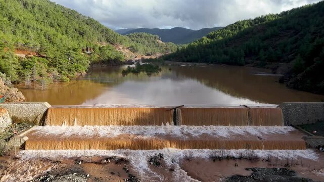 Muddy floodwaters of the Namnam River surging over a concrete stepped weir surrounded by pine-forested hills in Koycegiz, Mugla, Turkey. Aerial drone view, with moving forward