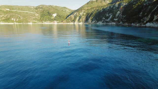 Man paddleboarder drifts across calm waters near forested hills of Kefalonia Island