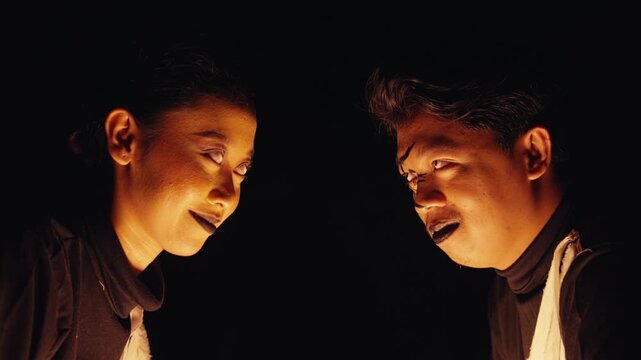 A man and woman with creepy makeup and white contacts face each other in darkness, lit from below. They share a menacing, conspiratorial look, creating a tense and evil atmosphere.