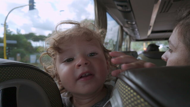 Toddler peeks between bus seats with playful expression, smiling slightly while wedged in small space during public transportation ride