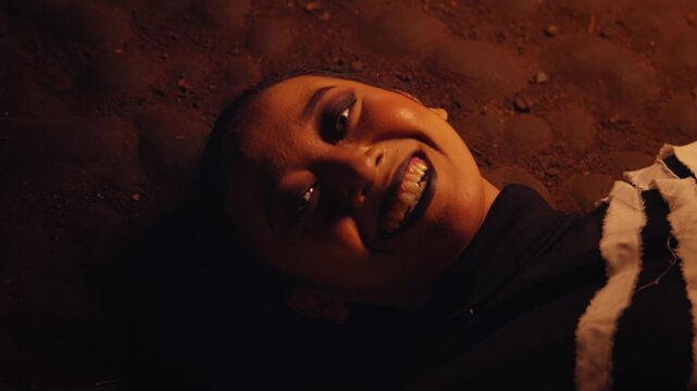 From a top-down view, a woman with theatrical makeup and white eyes lies on the ground in the dark. She slowly forms a sinister, evil smile, looking directly at the camera.