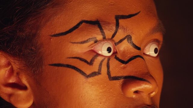 An intense extreme close-up focuses on a person's eyes, decorated with spider makeup and white contacts. The eyes are wide with fear, darting nervously in a state of panic or paranoia.
