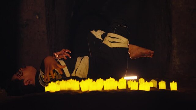 A man in a dark costume with theatrical makeup performs an interpretive dance while lying on the ground, surrounded by a line of candles. A strange and spiritual performance art piece.