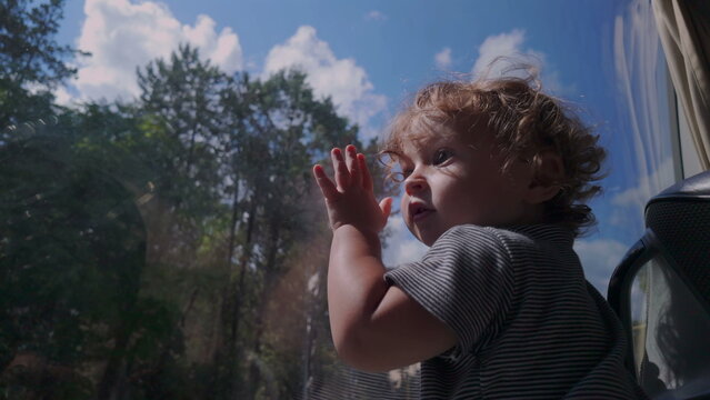 Toddler presses hand and face against bus window while observing outdoors, immersed in changing scenery with blue sky and trees reflected on glass