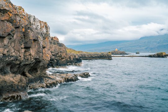 View of rugged basalt cliffs and ocean waves crashing against the shore with a small orange lighthouse in the distance under a cloudy sky Djupivogur, Mulathing, Iceland.