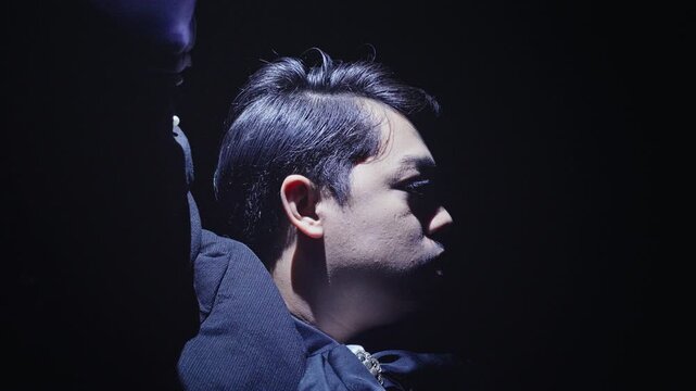 A dramatic close-up of a man's face in profile, illuminated by a harsh light against a black background. He raises his arm, creating a powerful and mysterious silhouette.
