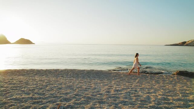 Woman walks barefoot along the tranquil pebble beach at sunset on Kefalonia island in Greece