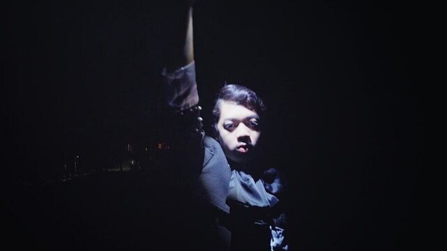 A man in a stylish outfit stands alone in the dark, using a flashlight to create dramatic lighting on his face before the scene fades into a wide, mysterious shot of him exploring.