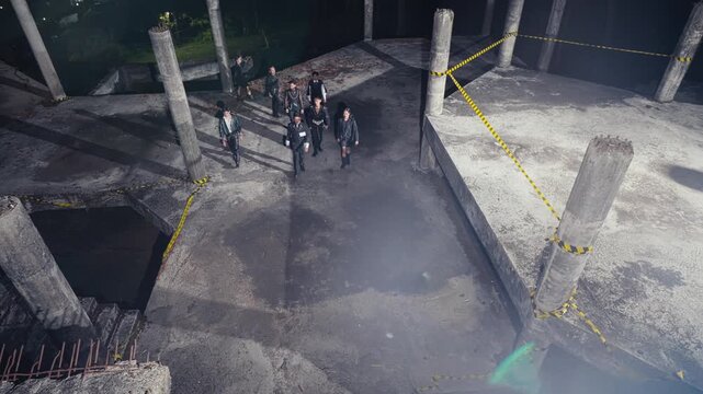 Seen from a high angle, a group of people dressed in black walks towards the camera through a derelict concrete structure, creating an intimidating and suspenseful atmosphere.