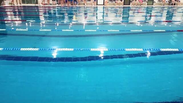 Swimmers at the edge of the indoor pool. Swimming competition in front of an empty pool. Blue water, dividing lines, red and white ropes, tiled flooring.