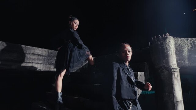 A man and woman dressed in edgy black fashion pose on a concrete staircase in a dark, abandoned location. The camera pulls out from a close-up to a wide shot, emphasizing their cool attitude.