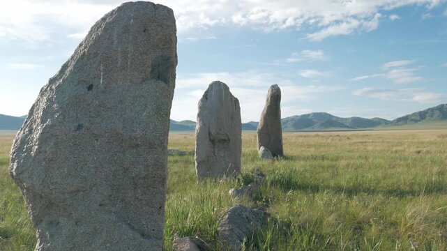 Handheld static shot of ancient kurgan standing stones in the vast grassy steppe of Khakassia. The historical megaliths stand tall against a beautiful background of green hills and a blue sky.