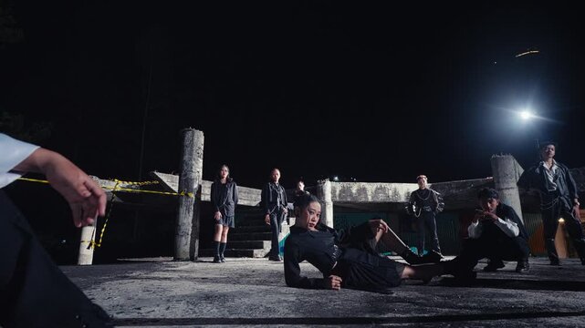 In a derelict building at night, a group of young models in stylish black attire hold dramatic poses. A low-angle shot with a person's arm in the foreground, creating an edgy, candid feel.