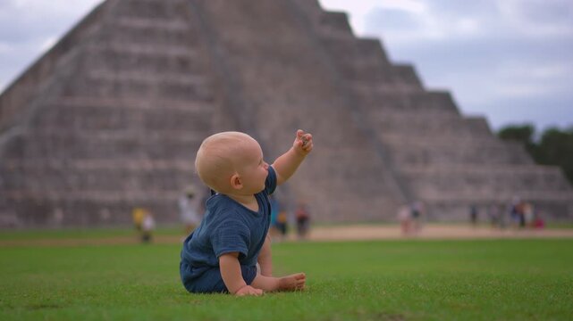 Cute one year old boy sitting and playing on grass in front of Kukulkan pyramid in Chichen Itza, Mexico. Happy child enjoying travel outdoors.