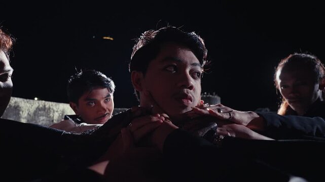 A young man is held captive or adored by his followers, who gently hold his face and neck. The scene is shot at night with a dramatic backlight, creating a tense, ritualistic mood.