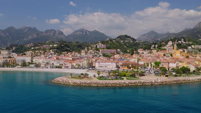 Aerial view of Menton on the French Riviera shows pastel old town, yellow church bell tower, curved breakwater, beach, marina with yachts, palms, and Maritime Alps.
