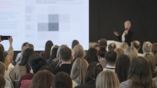 Business professionals in conference audience listening to presenter, taking photos, engaging with slides on screen.
