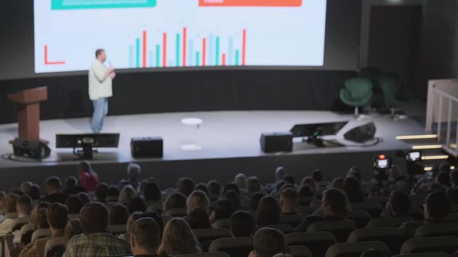Speaker presents data slides on stage at tech conference, audience seated in auditorium, camera recording.