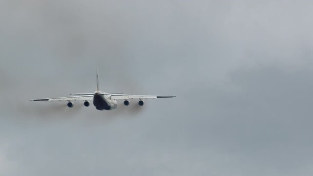 Massive white four engine cargo aircraft seen from rear in level flight, overcast gray sky, wide wingspan, engines clearly visible below wing
