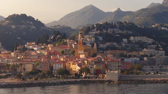 Aerial Menton, France, with Basilica of Saint Michel, cypress hills, Alpine ridges. Camera sweeps waterfront, breakwater, fort tower, promenade, boats, warm golden hour.