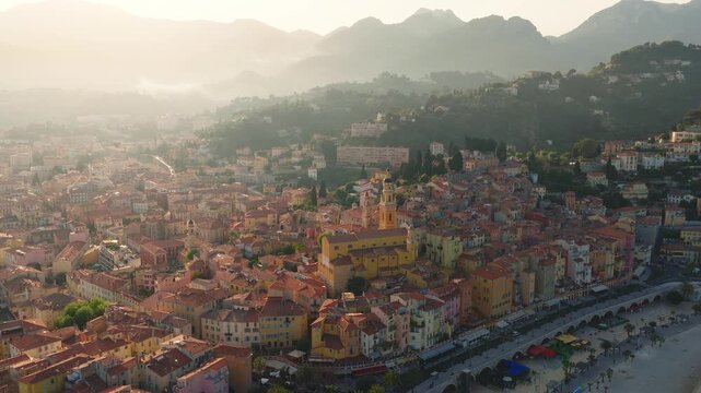 Aerial drone pans over Menton, French Riviera, showing crescent bay, pebbled beach, seafront arcades, market canopies, Basilique Saint Michel, docks, and calm sea.
