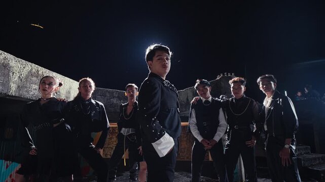 A stylishly dressed group of young people stands together in a powerful formation on a derelict rooftop at night. The leader in the center looks confidently at the camera, backlit by a bright light.