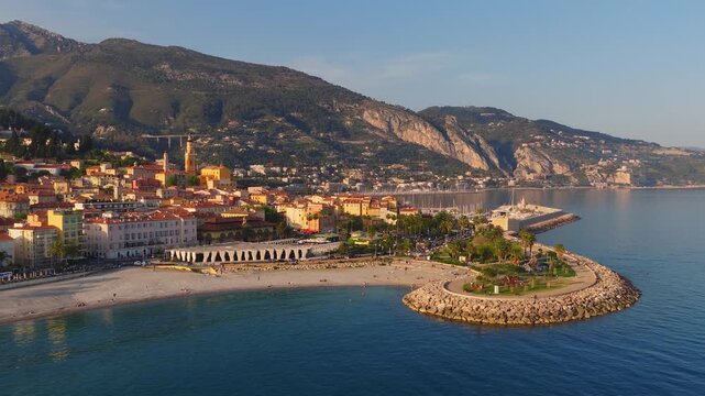 Aerial view of Menton, France shows old town pastel buildings, church bell tower, marina masts, curved stone breakwater, pebble beach, swimmers, and palm lined promenade.