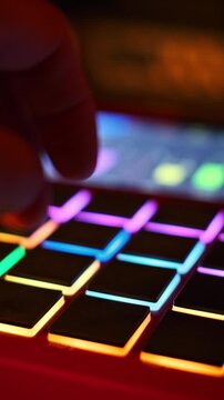 A musician finger taps the illuminated RGB pads of a music production controller in a dark studio. Concept of electronic music creation, nightlife entertainment, and digital beat making.