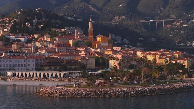 Aerial over Menton on the French Riviera shows pastel houses, ochre bell tower, curved breakwater, sandy beach, palm promenade, viaduct and hills in warm light.