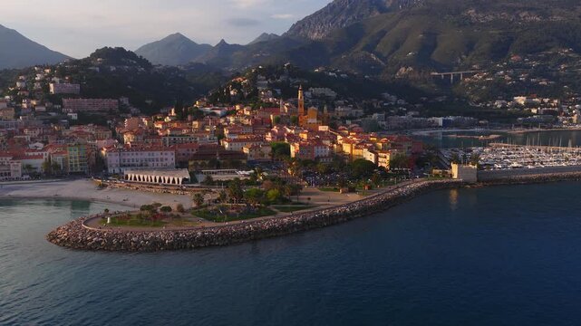 Aerial view of Menton, France old town and bell tower, curved breakwater, small fort, busy yacht marina, palm promenade and pebble beach in golden hour with motion